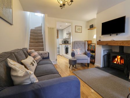 A living room with a grey sofa pillows an armchair a fireplace with a fire a wall mounted TV a bookshelf and stairs leading to another floor at Old Ropemakers Cottage in Bridport