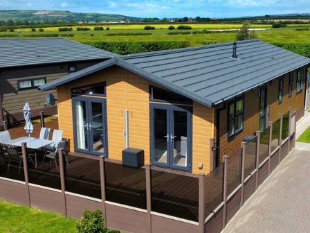 A modern wooden lodge with glass doors and fenced patio with outdoor dining set at Lodge 18 - The Pines in Runswick Bay