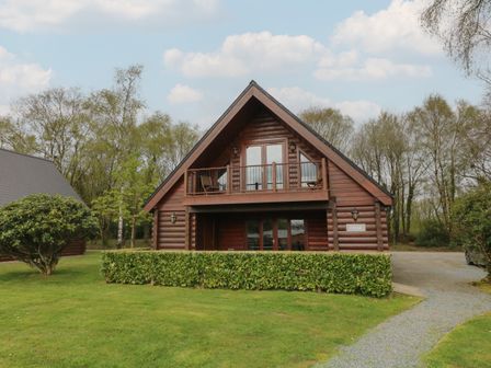 A wooden log cabin with a balcony and hedge in front surrounded by trees at Cedar in Dolton