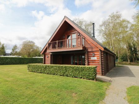 A wooden cabin with a balcony and hedge in front surrounded by grass and trees at Pine in Dolton