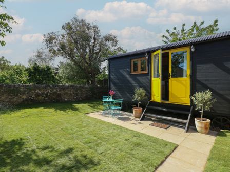 A small black cabin with yellow double doors on a stone patio with potted plants and a turquoise table and chairs on a grass lawn at Tower Hut in Ryde