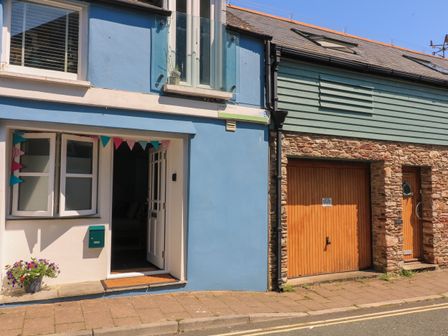A street with a blue house with an open door and a small garden pot next to it and a stone building with a wooden garage door and a wooden entrance door