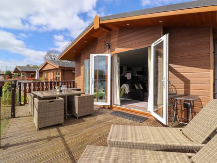 An outdoor wooden deck with wicker dining set and lounge chairs outside a wood-paneled house with open double doors showing interior at Capenwray in Carnforth