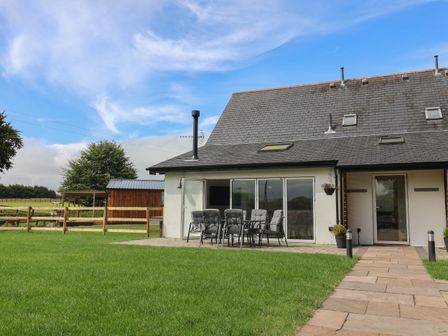 A house with a slate roof and glass doors opening to a patio with outdoor chairs and table with a grassy lawn at Campion Cottage in Brendon Hill near Roadwater