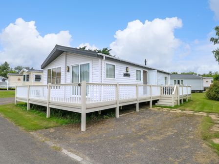 A white mobile home with a wraparound deck and steps on a grassy plot at 21 Norman Drive Lodge in Burgh Castle