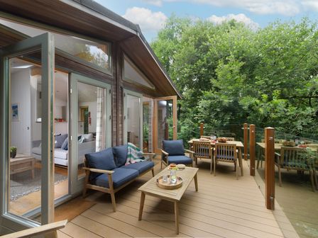 A wooden deck with blue cushioned chairs a wooden table and glass railing surrounded by trees at 10 Stonerush Valley Lanreath near Looe
