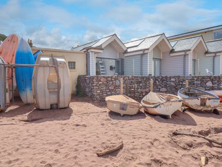 Small boats and surfboards on sandy ground in front of stone wall and light blue beach huts at Beach Hut 5 in Shaldon