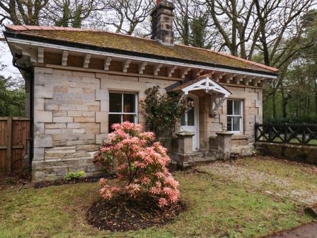 A small stone house with two windows and a white door with a porch a flowering shrub in front and trees in the background at Gatekeepers in Loftus near Easington