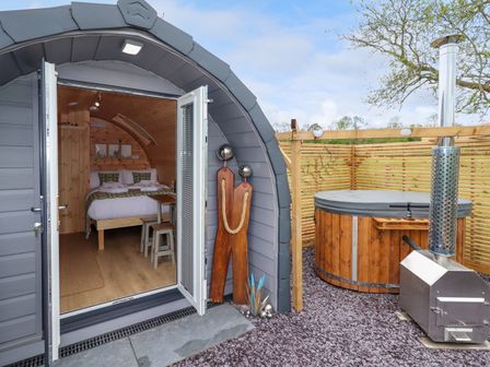 A small wooden pod with open doors showing a bed inside and a covered hot tub with heater outside at Moel Arthur in Bryniau Clwyd near Denbigh