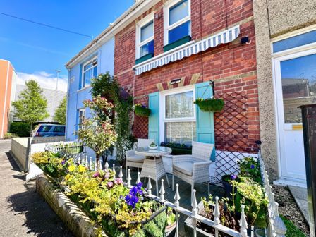 A small outdoor seating area with chairs and a table surrounded by flowers in front of a brick house with blue shutters at Garibaldi Cottage in Weymouth