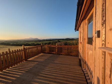 A wooden deck with a picket fence attached to a wooden cabin overlooking fields at Hafan Gelli Sheperds Hut - Tudwal in Pwllheli