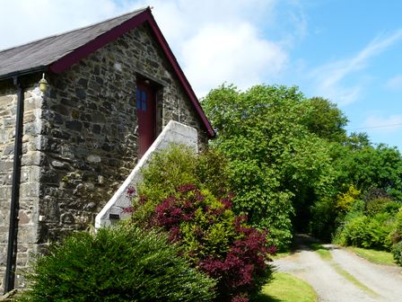 A stone house with an external staircase and a red door surrounded by bushes near a gravel driveway