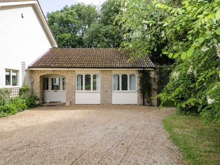 A small stone cottage with white windows and a door surrounded by trees and greenery at Garden Cottage in Acton Turville