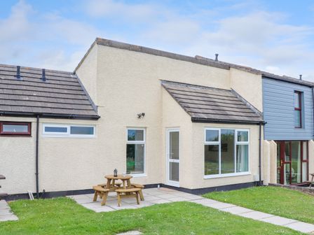 An exterior view of a beige house with a small patio area with a round wooden picnic table and grassy lawn at Skylarks Trevellas Nr St Agnes