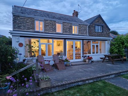 The exterior of a two story stone house with illuminated windows a patio with wooden chairs and a picnic table and flower pots at Langurra in Crantock