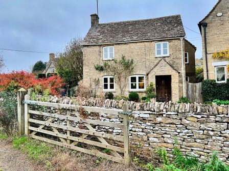 A stone house with white windows behind a wooden fence and stone wall at Meadow View Cottage in Calcot near Northleach