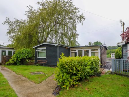 A small dark wooden house with white framed windows and a pathway leading to the entrance surrounded by greenery and trees at 245 Riverside Lodge in Caeathro