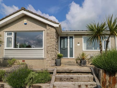 Front view of a single story house with two large windows and a double door entrance with steps and plants at Benllech Bay Cottage in Benllech