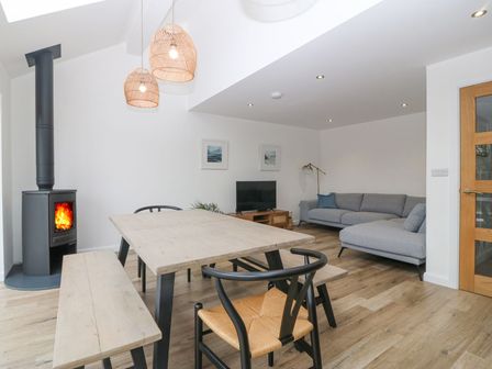 A dining and living area with wooden table bench chairs wood burning stove and grey sofa at 11 Capel Farm in Trearddur Bay