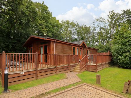 A wooden cabin with a surrounding deck and outdoor seating in a wooded area at Templar Rise 16 - Devon Comfort Finlakes Holiday Park near Chudleigh