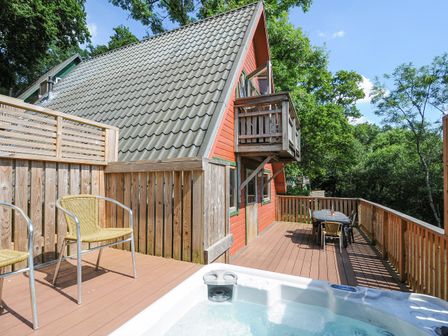 An outdoor deck with a hot tub two chairs a dining table and an A-frame house surrounded by trees at Lodge 47 - Primrose Lodge in Finlakes Holiday Park near Chudleigh