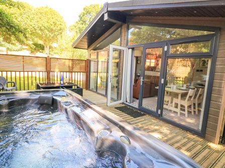 An outdoor deck with a hot tub a table with chairs and a dining area visible through glass doors at Woodland View 7 - Oak Cabin Finlakes Holiday Park near Chudleigh