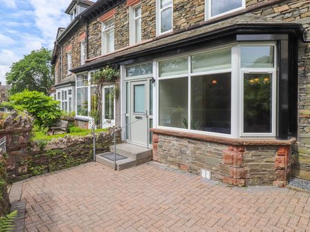 Front view of a stone house with a white door and large windows a garden area with plants and a bench at Eller Cottage in Windermere