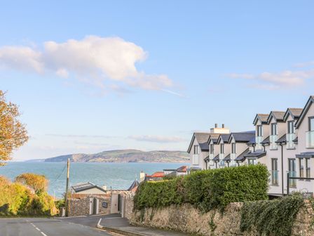 A coastal road with houses on the right side and a view of the sea and hills in the background at Sandy Bay Retreat in Benllech