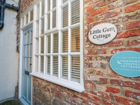 A blue door and white window on a brick wall with signs for Little Gem Cottage in Whitby