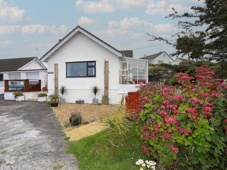 A white house with a driveway and garden with pink flowers and plants at Greenfield in Trearddur Bay