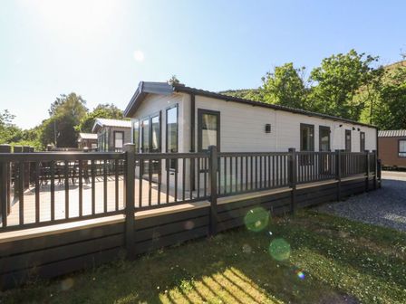 An exterior view of a white mobile home with a wooden deck and black railing surrounded by grass and trees at Saorsa in St Fillans