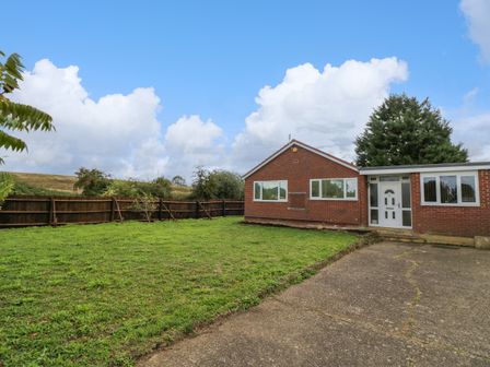 A single-story brick house with a lawn and driveway under a blue sky at Fenny Marina Cottage in Fenny Bentley