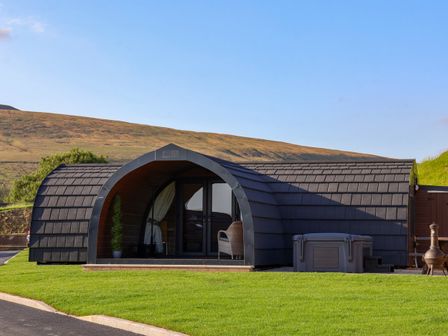A curved black cabin with glass doors and a wicker chair outside on green grass at Heron in Ingleton