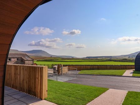 A view of green fields and distant hills from a curved wooden structure with a fenced yard at Wren in Ingleton
