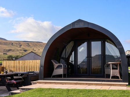 A curved pod structure with two wicker chairs on the porch next to a picnic table with a bottle and glasses at Barn Owl in Chapel Le Dale near Ingleton