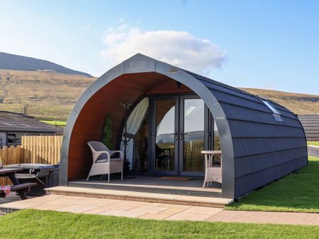 A curved wooden pod with glass doors, two wicker chairs, and a small table on the porch at Curlew in Chapel Le Dale near Ingleton