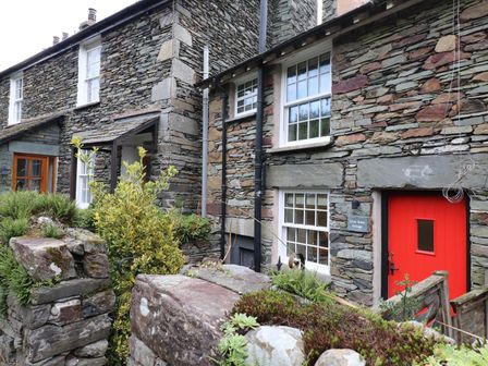 The exterior of a stone cottage with white framed windows and a red door surrounded by plants and a stone wall at Little Robin Cottage in Ambleside