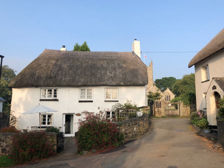 A thatched roof cottage with white walls and outdoor seating on a street near a stone church at Primrose Cottage in Drewsteignton