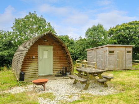 A wooden outdoor pod with a green door beside a wooden shed and a picnic table on gravel at Rabbit Warren Penbugle Organic Farm in St Keyne near Duloe