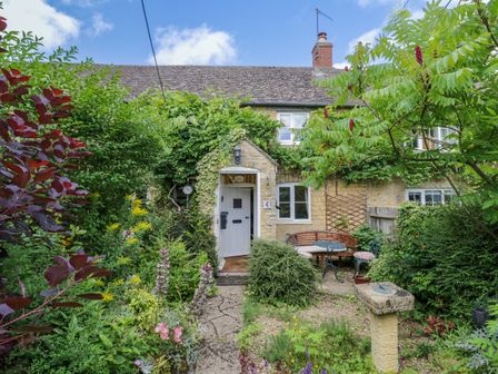 A stone cottage with a white door surrounded by greenery and a garden with outdoor seating at 4 Lansdown Cottages Bourton-On-The-Water