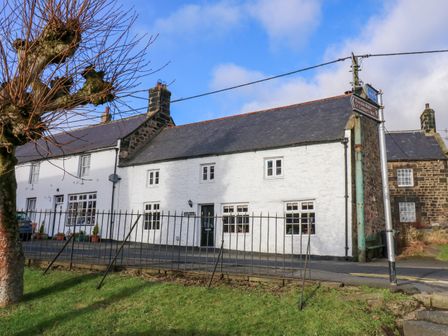 A row of traditional stone cottages with white and stone exterior walls next to a road and a metal fence in front at Rose Hip Cottage in Bellingham