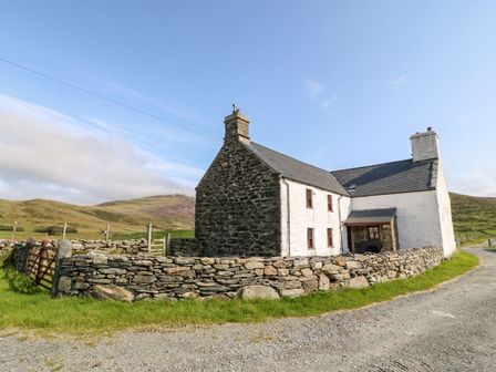 A stone and white cottage with a slate roof surrounded by a low stone wall on a gravel road in a rural area at Ereiniog 2 in Tremadog