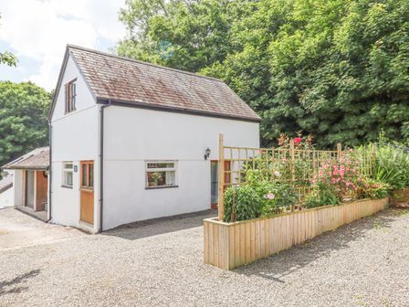 A white two-story building with a gravel driveway and a wooden planter with flowers and trellis at Dove Cottage Queens Nympton near South Molton