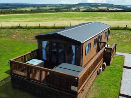 A wooden cabin with a deck and glass doors surrounded by grass and fields at Hazel Lodge in Whitby