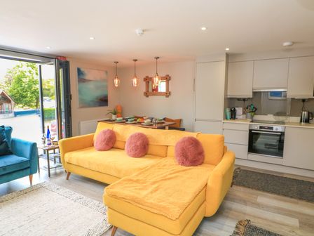 An open living area with a yellow sofa, dining table with pendant lights, kitchen cabinetry, and an open door to outside at 2 De Courcey House in Dartmouth