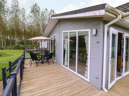 A wooden deck with a table and chairs under an umbrella outside a house with large windows and a glass door at Owls Retreat Lodge 11 Near Aldeburgh