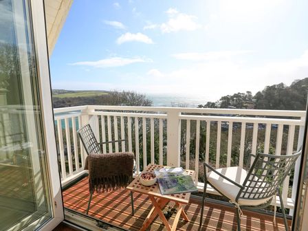 A balcony with two chairs a small table and a view of trees and the sea at 6 Grafton Towers in Salcombe