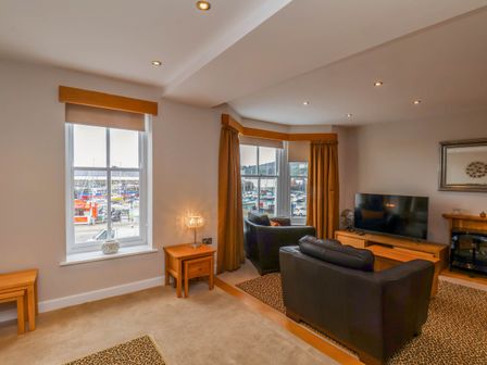 A living room with black leather sofas a wooden TV stand and bay windows overlooking a marina at 43A Quay Street in Scarborough