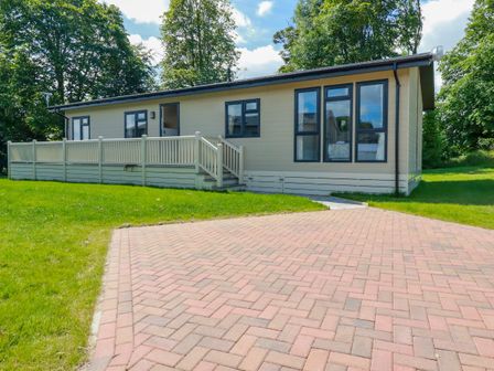 A beige single-story mobile home with windows and a small porch on a lawn with a paved driveway at 43 The Maples in Sticker