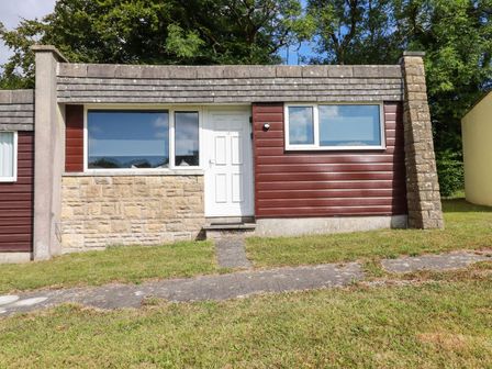 A single story building with wooden paneling stone facade white door and large windows on grass at Villa 18 Lanteglos Holiday Park near Camelford